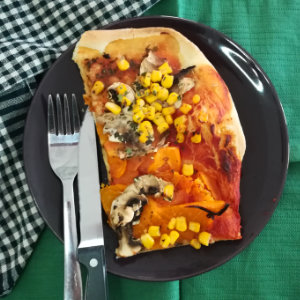 Vegane Pizza mit Tomaten, Pilzen und Mais belegt auf einem Teller mit Messer und Gabel