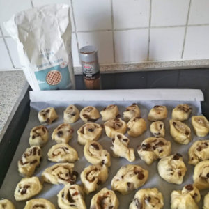 Vegane Rosinen-Zimtschnecken auf einem Backblech. Im Hintergrund befinden sich eine Packung Zucker und Zimt